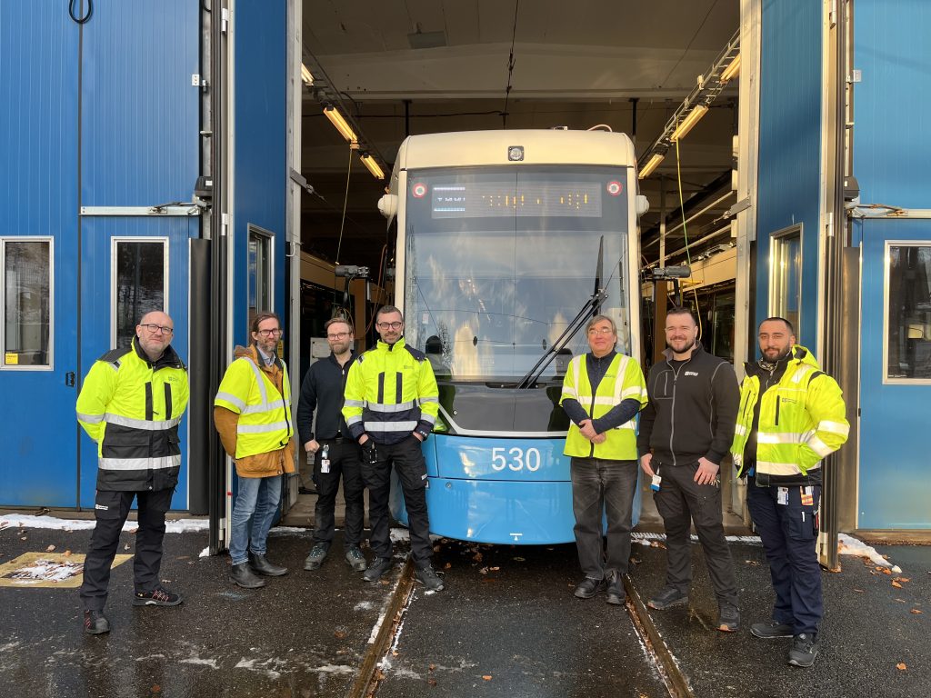Sju personer står utomhus framför fronten på en spårvagn som är utkörd genom två blå portar.